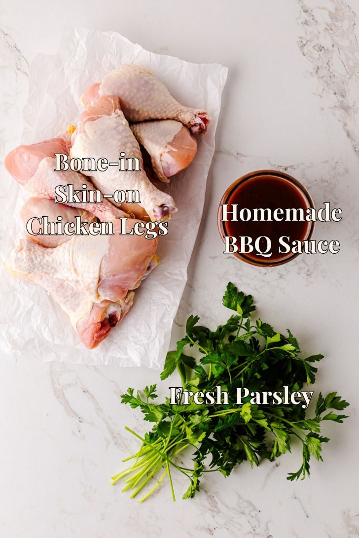 Overhead view of raw bone-in, skin-on chicken legs on parchment paper, a bowl of homemade BBQ sauce, and a bunch of fresh parsley on a marble countertop, each labeled with text.