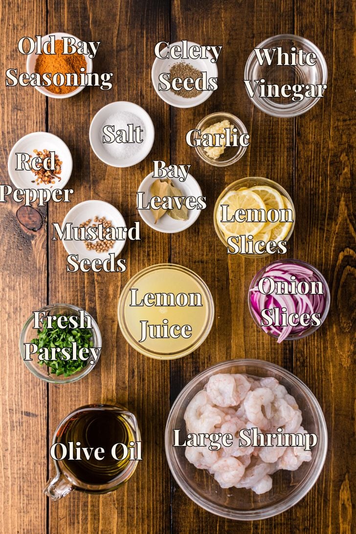 Overhead view of labeled ingredients for pickled shrimp arranged on a wooden surface, including large shrimp, olive oil, lemon juice, white vinegar, lemon slices, onion slices, garlic, fresh parsley, Old Bay seasoning, celery seeds, mustard seeds, bay leaves, red pepper flakes, and salt.