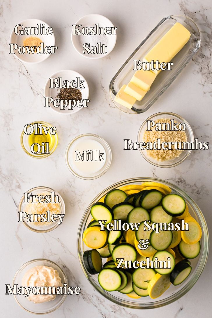 Overhead view of labeled ingredients for zucchini and squash casserole on a marble surface, including sliced yellow squash and zucchini, butter, panko breadcrumbs, mayonnaise, milk, olive oil, garlic powder, kosher salt, black pepper, and fresh parsley.