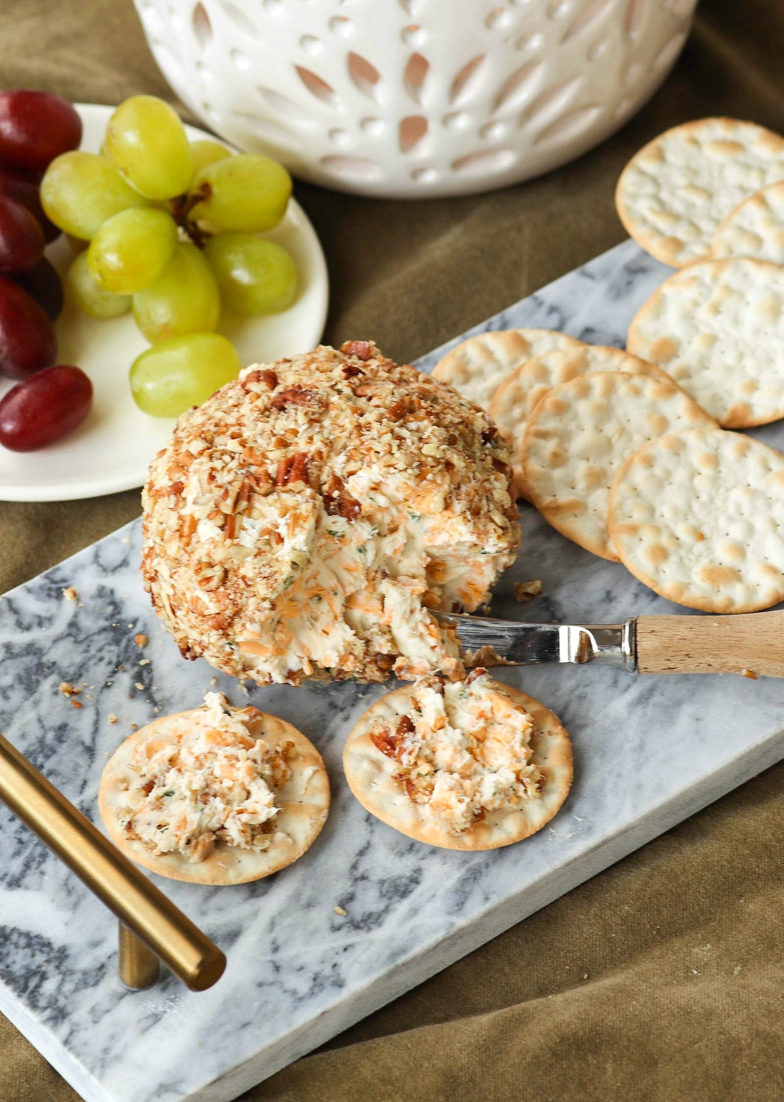 Pecan-crusted cheese ball spread on crackers with grapes on a marble board.