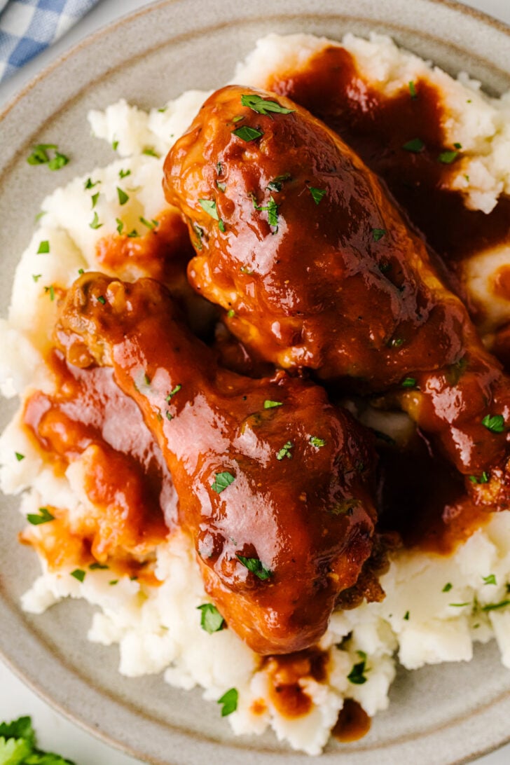Close-up of two BBQ-sauced chicken legs served over a bed of creamy mashed potatoes on a gray plate, garnished with chopped parsley.