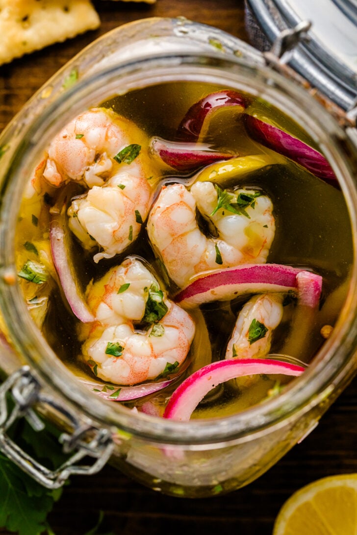 Overhead close-up of a glass jar filled with pickled shrimp, red onion slices, fresh herbs, and lemon in a seasoned brine. The shrimp are plump and marinated, surrounded by vibrant pickling ingredients.