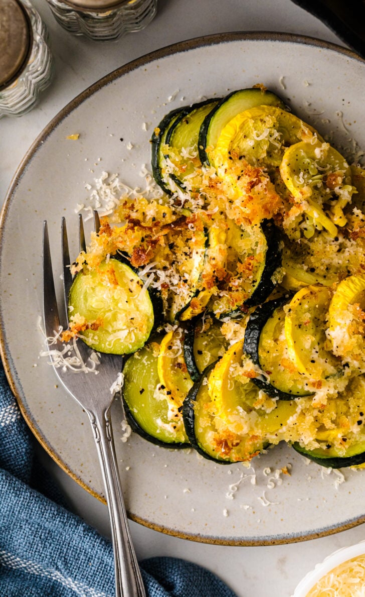Plate of baked zucchini and yellow squash casserole topped with golden, crispy breadcrumbs and shredded cheese, served with a fork on a ceramic plate.