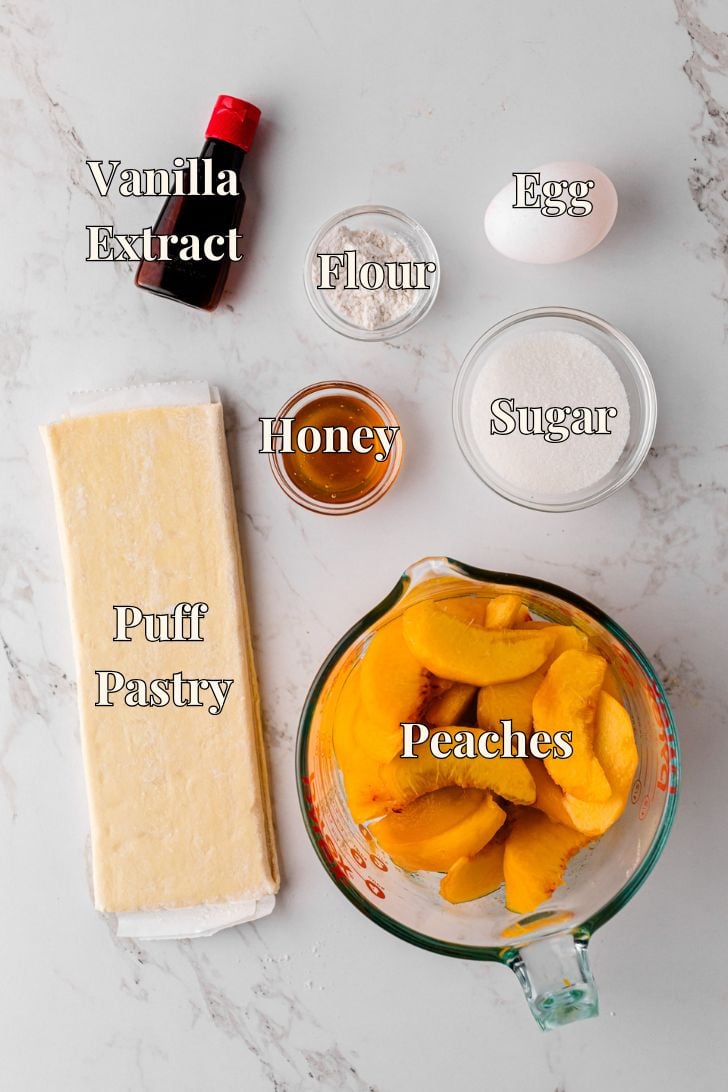 Flat lay of peach tart ingredients on a marble surface, including sliced peaches in a measuring cup, puff pastry sheet, honey, sugar, flour, egg, vanilla extract, all clearly labeled. The setup is bright and clean, emphasizing each component of the dessert.