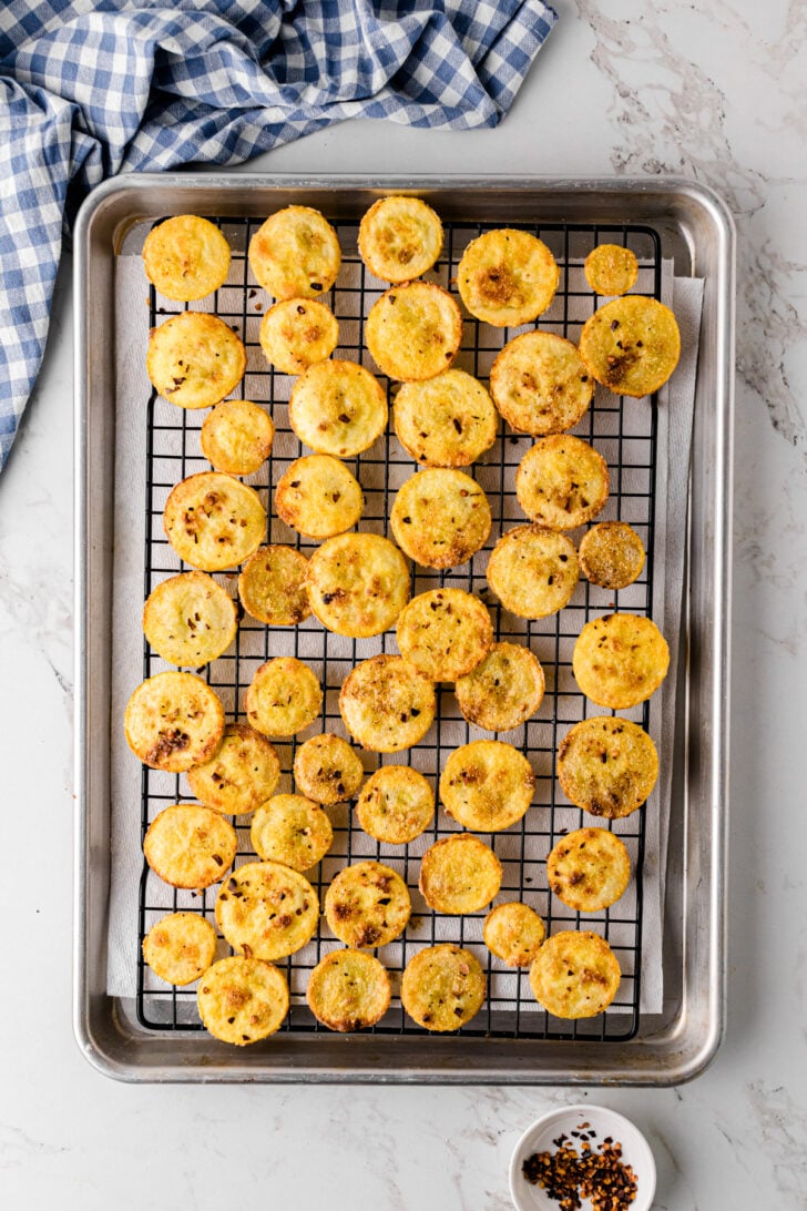 Golden brown fried squash rounds cooling on a wire rack set over a baking sheet lined with paper towels. A blue gingham towel sits in the upper left corner, and a small bowl of red pepper flakes is in the lower right.