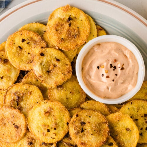 Plate of golden fried squash with crispy edges and black pepper seasoning, served with creamy tan dipping sauce on white dish