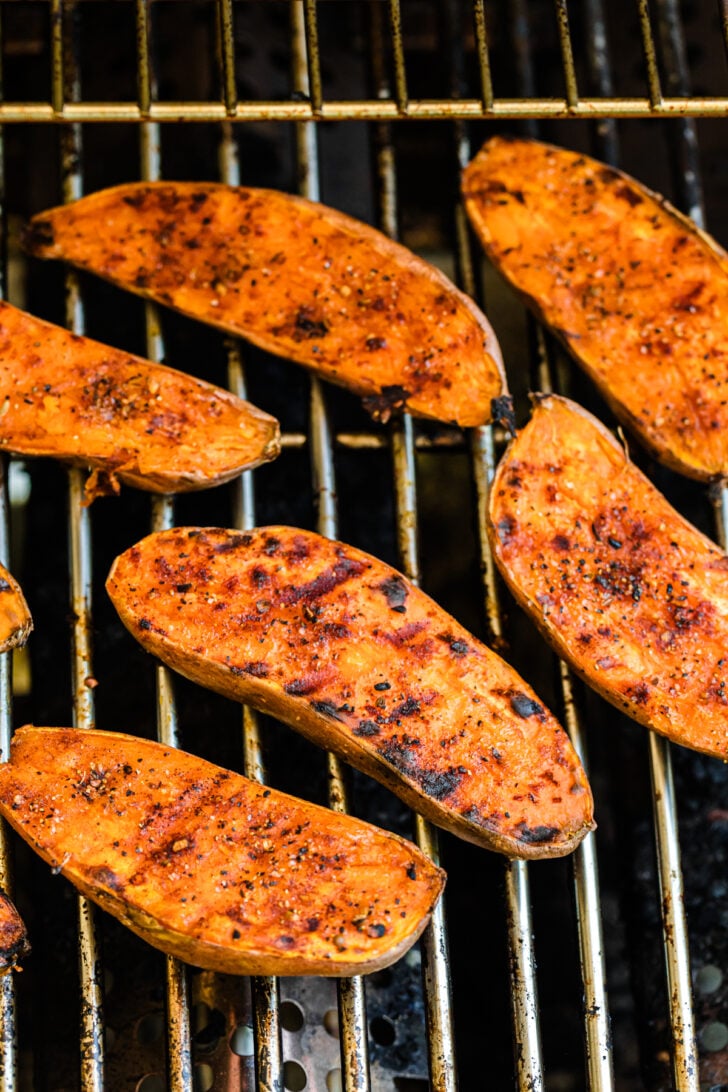 Sweet potato halves grilling over open flames, with visible char marks and seasoning. The grill grates are slightly worn, adding a rustic outdoor cooking feel. The potatoes have a vibrant orange color and lightly caramelized edges.