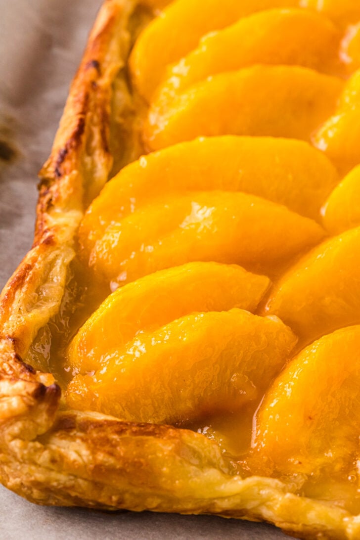 Golden-brown puff pastry peach tart with glistening peach slices arranged in rows, showcasing flaky crust edges and glossy fruit topping. The tart rests on parchment paper for a rustic presentation.