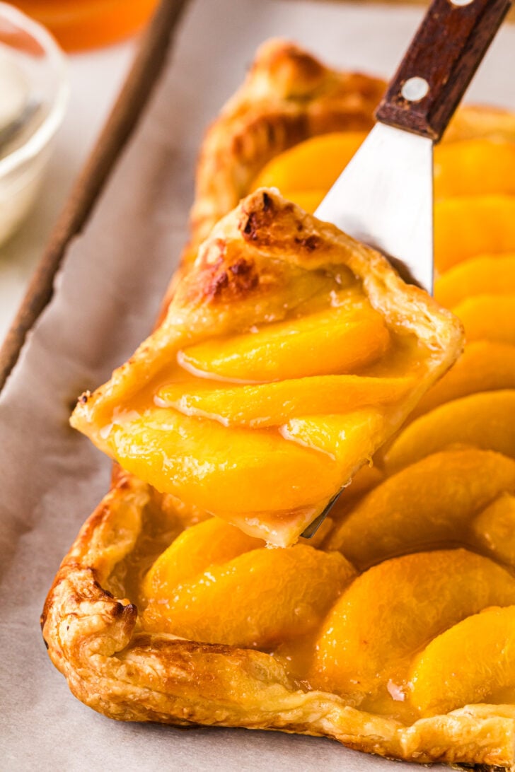 A golden slice of peach tart being lifted from the baking sheet with a metal spatula. The flaky puff pastry crust is crisp and browned, and the tart is layered with glossy, vibrant peach slices in a rich glaze.
