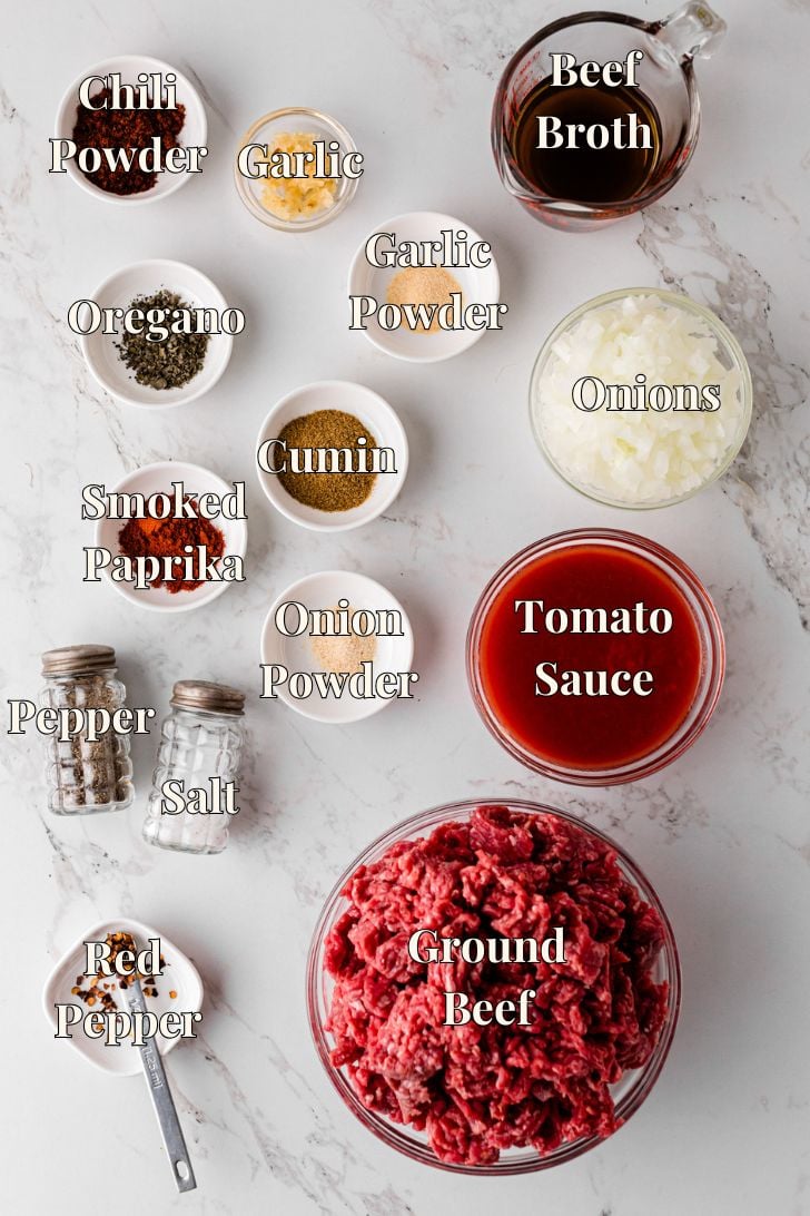 Overhead view of chili seasoning and meat ingredients arranged on a white marble surface. Ingredients include bowls of chili powder, minced garlic, garlic powder, oregano, cumin, smoked paprika, onion powder, red pepper flakes, chopped onions, ground beef, tomato sauce, and beef broth. Salt and pepper shakers are also shown