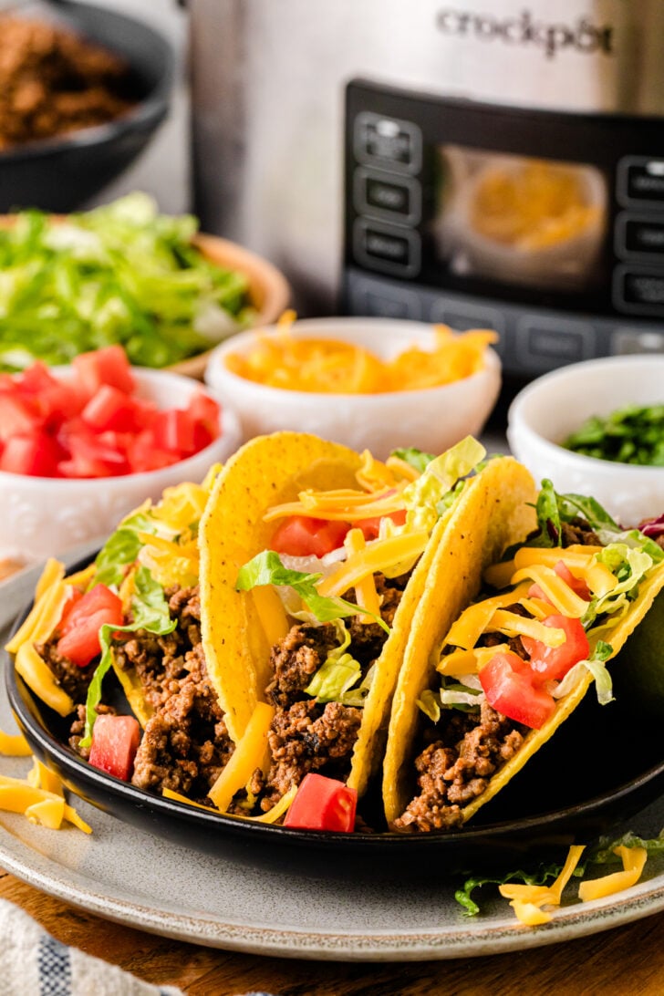 Plate of crunchy hard-shell beef tacos loaded with seasoned ground beef, shredded lettuce, diced tomatoes, and cheddar, with a slow cooker and taco toppings in the background.