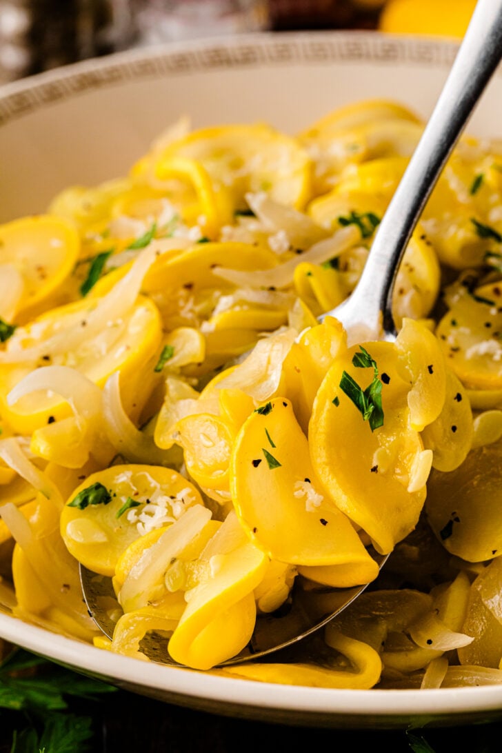 A close-up view of a spoon lifting tender slices of sautéed yellow squash and onions from a serving bowl. The vegetables are garnished with chopped parsley, black pepper, and a sprinkle of salt, highlighting their glossy, buttery texture.