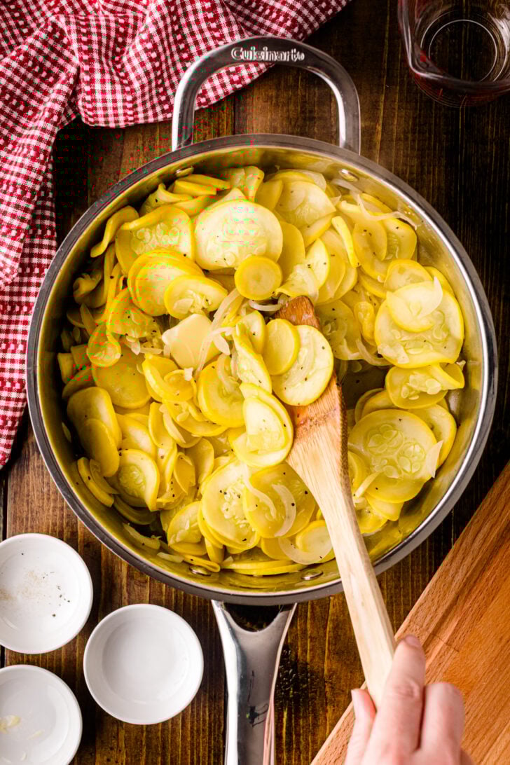 A hand stirs a skillet of sautéed yellow squash and onions with a wooden spoon. The vegetables are lightly seasoned and glistening, while a red and white checkered towel and small empty bowls rest on the rustic wood counter nearby.