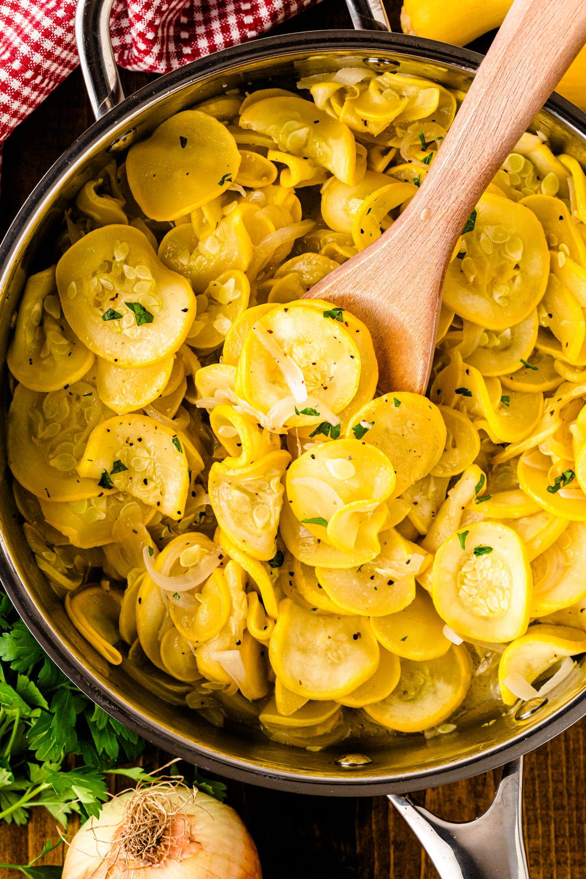 A stainless steel skillet filled with tender slices of yellow squash and translucent sautéed onions, garnished with cracked black pepper and fresh parsley. A wooden spoon lifts a portion from the pan, and a red gingham towel and sweet onion sit nearby on the rustic wooden table.