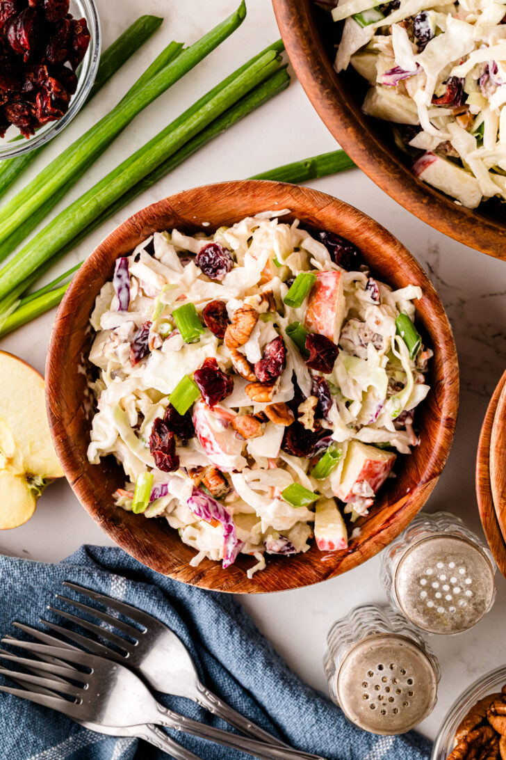 Overhead view of a wooden bowl filled with creamy coleslaw made with cabbage, apples, dried cranberries, pecans, and green onions, surrounded by fresh ingredients and serving utensils.