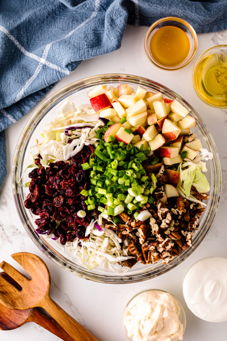 Overhead view of a glass bowl filled with chopped apples, shredded cabbage, dried cranberries, green onions, and pecans, ready to be mixed into a fresh salad. Small bowls of dressing and wooden salad servers sit nearby.