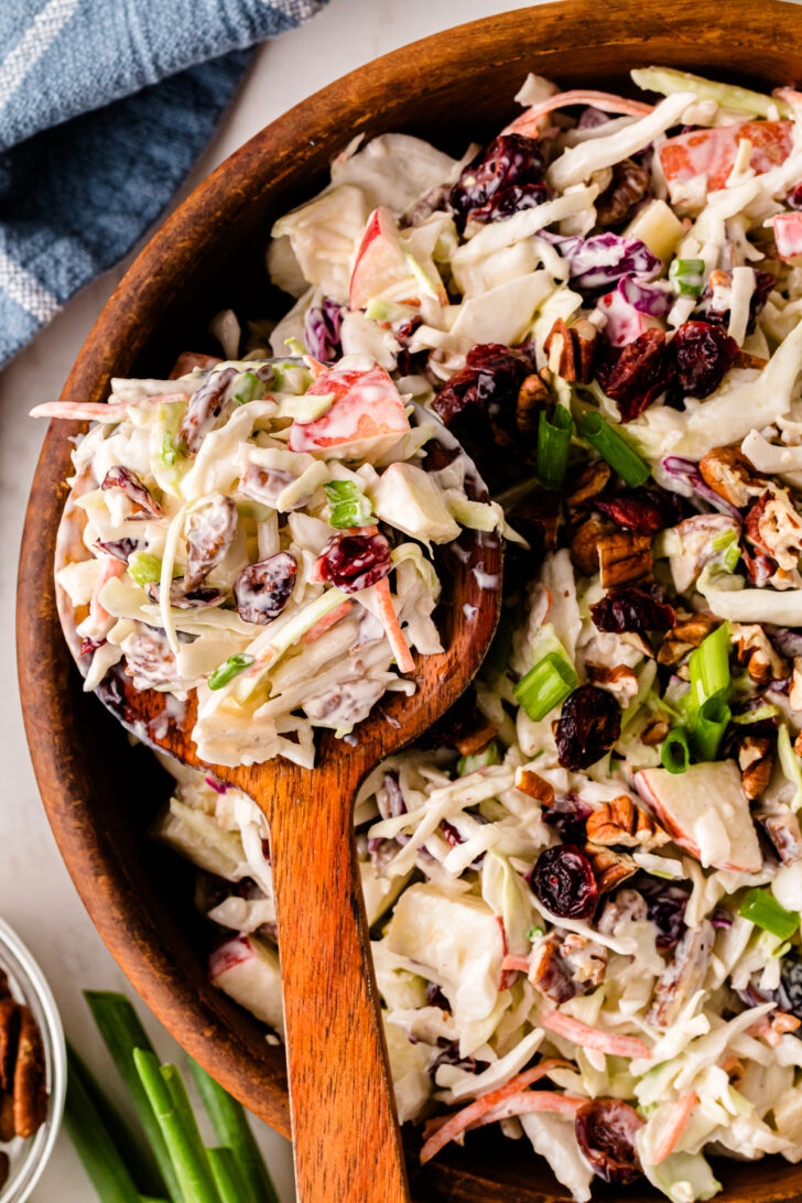 Wooden spoon scooping creamy coleslaw made with cabbage, apples, dried cranberries, pecans, and green onions from a wooden serving bowl.