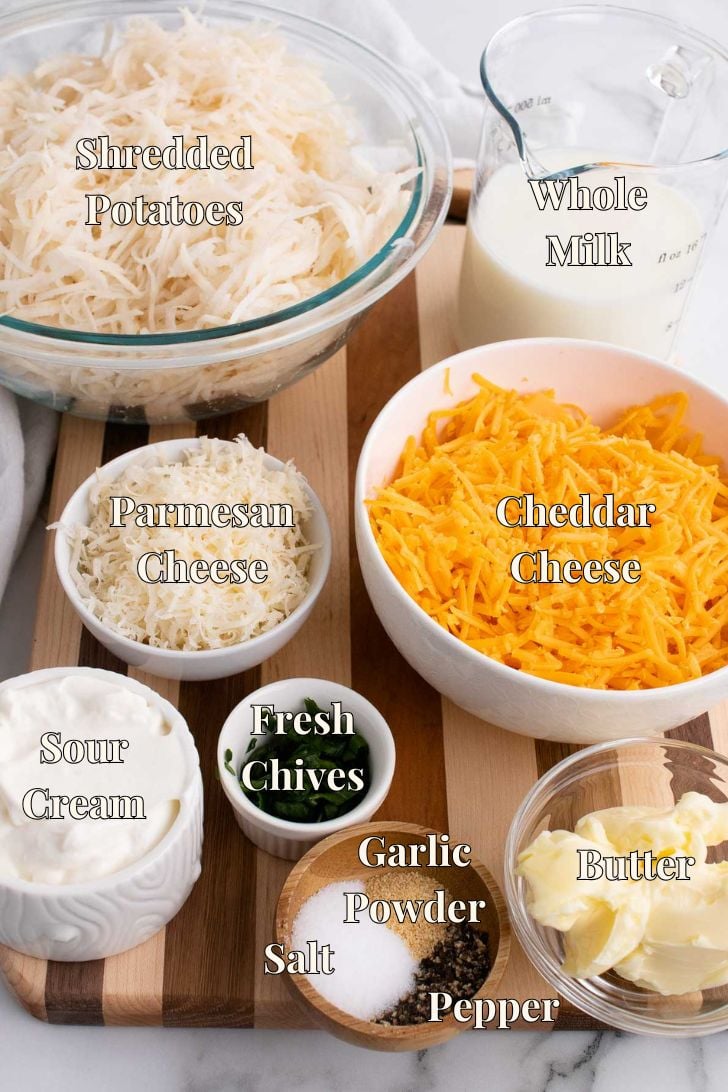 Overhead view of hashbrown casserole ingredients: shredded potatoes, whole milk, cheddar cheese, parmesan cheese, sour cream, fresh chives, butter, garlic powder, salt, and pepper arranged on a wooden board.