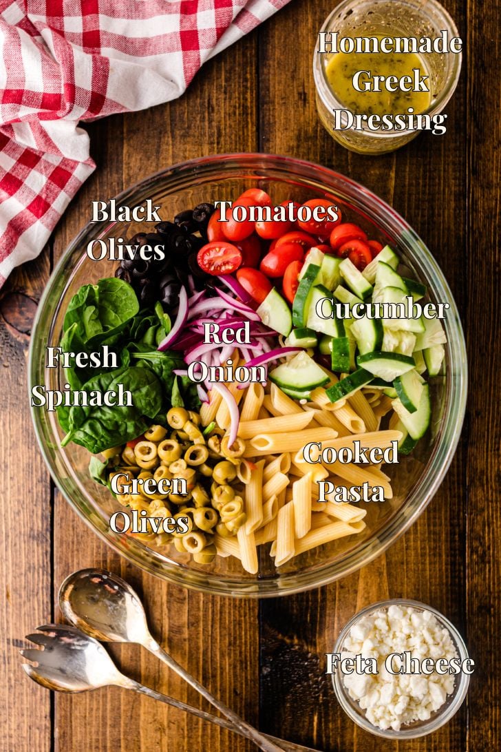 Overhead view of Mediterranean pasta salad ingredients in a glass bowl: cooked penne pasta, cucumbers, cherry tomatoes, red onion, black olives, green olives, and fresh spinach, with small bowls of feta cheese and homemade Greek dressing on the side.