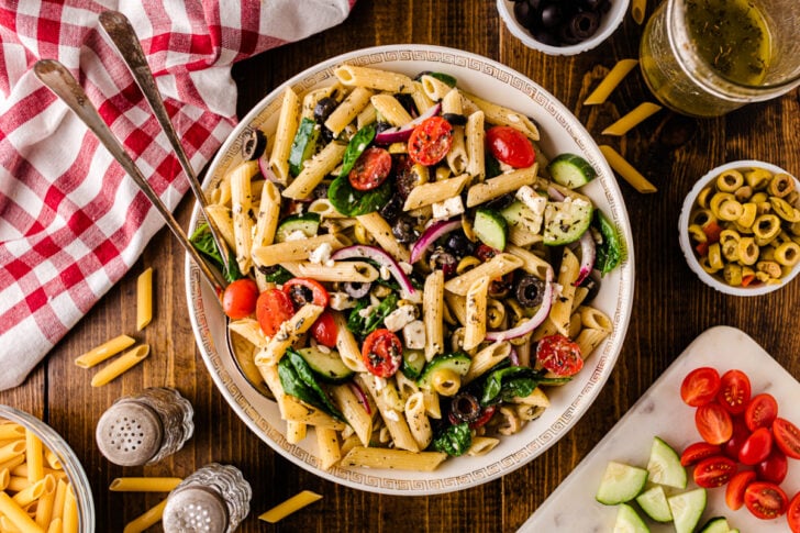 Large bowl of Mediterranean pasta salad with penne, cherry tomatoes, cucumbers, black olives, red onion, spinach, and feta, surrounded by fresh ingredients and a red checkered napkin on a wooden table.