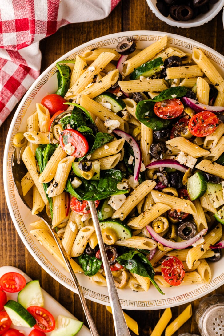 Overhead view of Mediterranean pasta salad with penne, cherry tomatoes, cucumbers, black olives, red onion, spinach, and feta cheese on a white plate, surrounded by extra ingredients and a red checkered napkin.