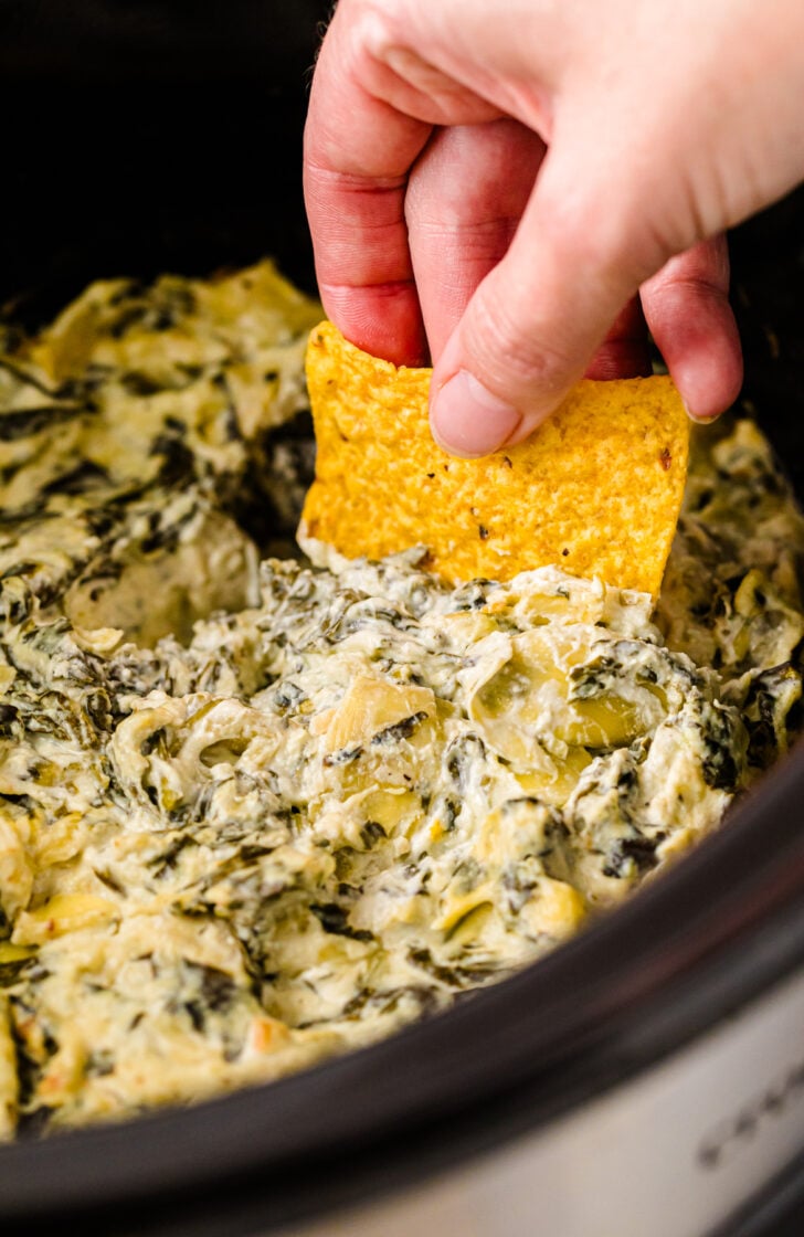 Close-up of a hand dipping a tortilla chip into creamy spinach artichoke dip inside a slow cooker
