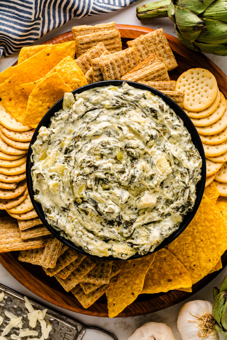 Overhead view of a bowl of creamy spinach artichoke dip surrounded by tortilla chips, woven wheat crackers, and round buttery crackers on a wooden serving tray.
