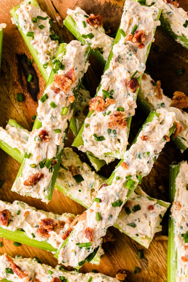 Overhead view of celery sticks stuffed with a creamy cheese mixture, topped with crumbled bacon and chives, arranged on a wooden serving board with bowls of extra toppings nearby.