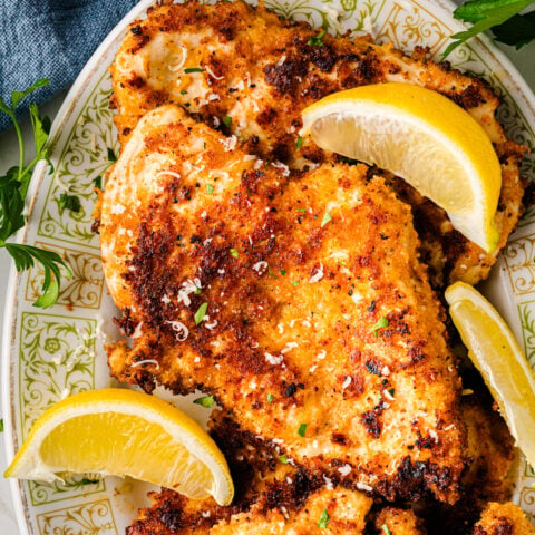 Parmesan crusted chicken breasts on decorative plate with lemon wedges and fresh parsley garnish, overhead view