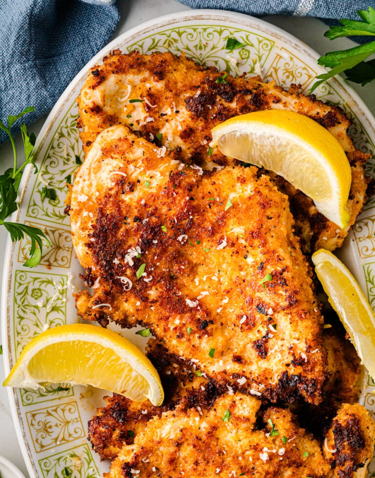 Parmesan crusted chicken breasts on decorative plate with lemon wedges and fresh parsley garnish, overhead view