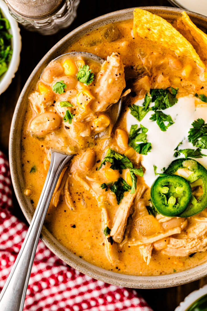 Close-up of a bowl of creamy white chicken chili topped with sour cream, sliced jalapeños, fresh cilantro, and tortilla chips, with a spoon lifting a serving of chicken, beans, and corn