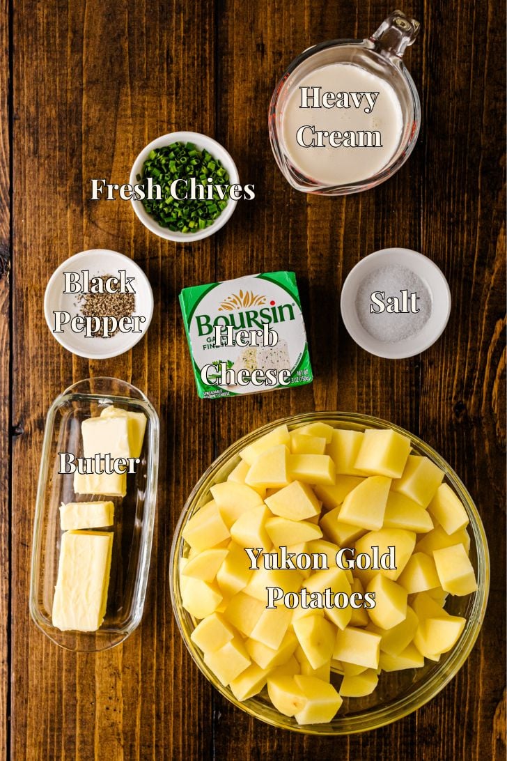 Overhead flat lay of recipe ingredients including cubed Yukon Gold potatoes, a package of Boursin garlic-herb cheese, butter, heavy cream, chopped chives, salt, and black pepper on a wooden board.