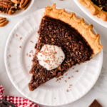 Chocolate chess pie slice on white plate topped with whipped cream and chocolate shavings, served with pecans and chocolate