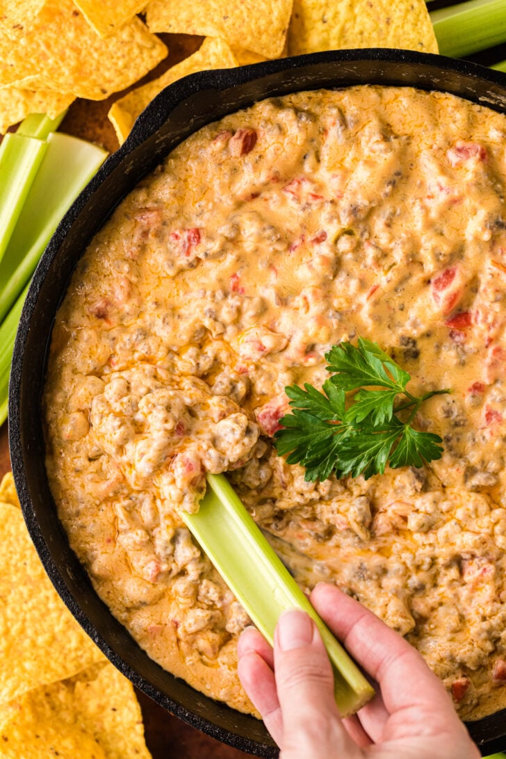 Hand dipping a celery stick into warm pimento cheese sausage dip, showing creamy texture and party-ready serving.