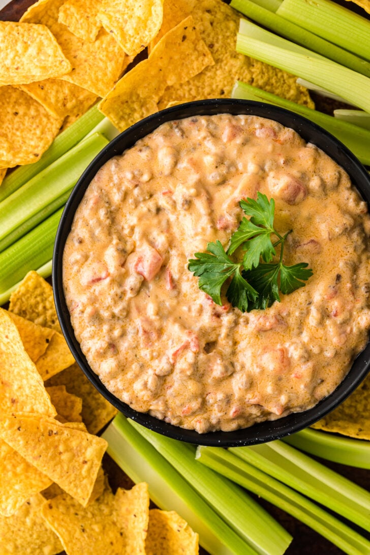 Pimento Cheese and Sausage Dip 23 in black bowl surrounded by tortilla chips and celery sticks, garnished with fresh parsley