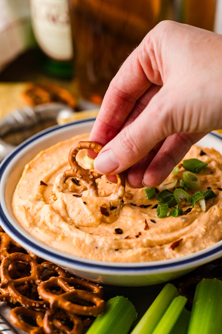 क्रॉक पॉट बियर चीज़ डिप एक सर्विंग बाउल में तैयार और मलाईदार है, ऊपर से हरा प्याज डाला गया है और डिपिंग के लिए प्रेट्ज़ेल, चिप्स और सब्जियों से घिरा हुआ है।