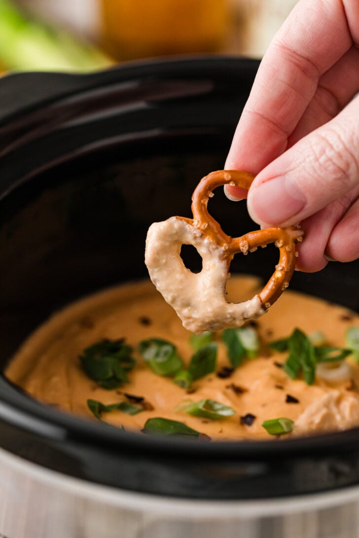 गर्म क्रॉक पॉट बियर चीज़ डिप में प्रेट्ज़ेल बाइट को हाथ से डुबाएं, जिससे प्रेट्ज़ेल को कोट करने पर इसकी गाढ़ी, मलाईदार बनावट दिखाई देती है।