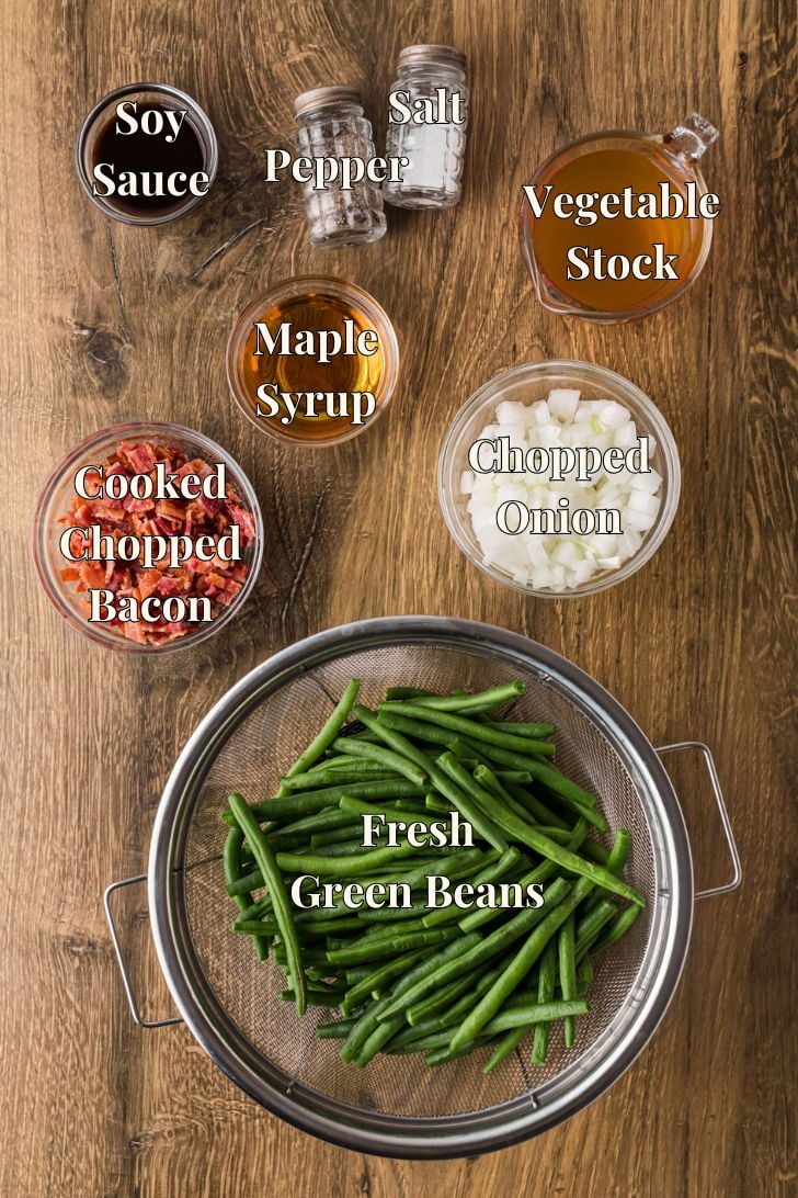 क्रॉक पॉट मेपल बेकन हरी बीन्स के लिए सामग्री एक काउंटर पर रखी गई है, जिसमें हरी बीन्स, बेकन, मेपल सिरप, शोरबा और मसाला शामिल हैं।