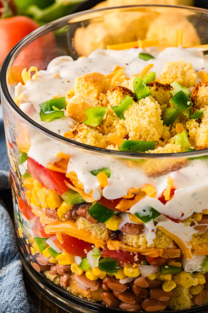 Overhead view of Southern Cornbread Salad in a glass trifle bowl topped with creamy ranch dressing, golden cornbread cubes, diced green bell pepper and shredded cheddar peeking through, set on a dark wood table beside a cast iron skillet of cornbread, fresh vine tomatoes, a green bell pepper, a blue and white checkered napkin and a black plate with forks