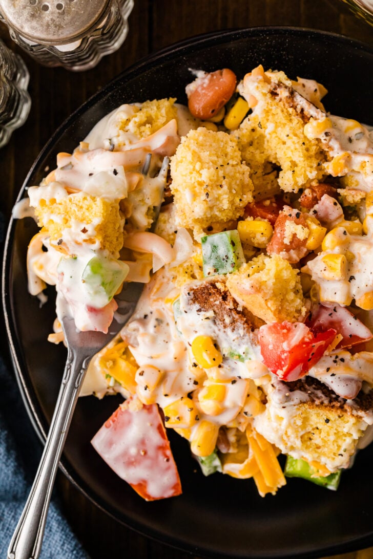 Overhead view of a single serving of Southern Cornbread Salad on a matte black plate with a vintage fork — tossed cornbread chunks, diced tomatoes, sweet corn, green bell pepper, shredded cheddar and creamy ranch dressing speckled with black pepper