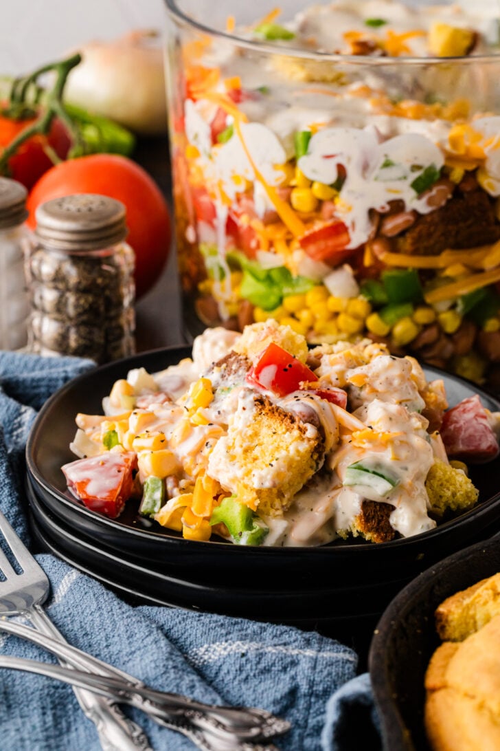 Black plate holding a creamy serving of Southern Cornbread Salad with cornbread cubes, diced tomatoes, corn, green pepper, cheddar and ranch dressing, with the layered glass trifle bowl, vine-ripe tomatoes, a yellow onion, a glass pepper grinder, blue denim napkin and antique silver forks styled in the background