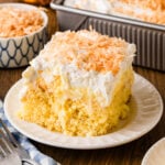 Pineapple poke cake slice topped with whipped cream and toasted coconut flakes on a white plate with a fork