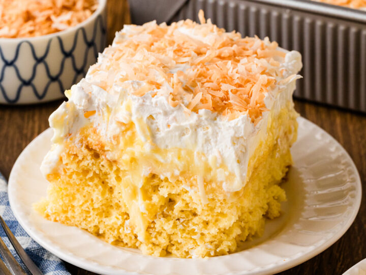 Pineapple poke cake slice topped with whipped cream and toasted coconut flakes on a white plate with a fork