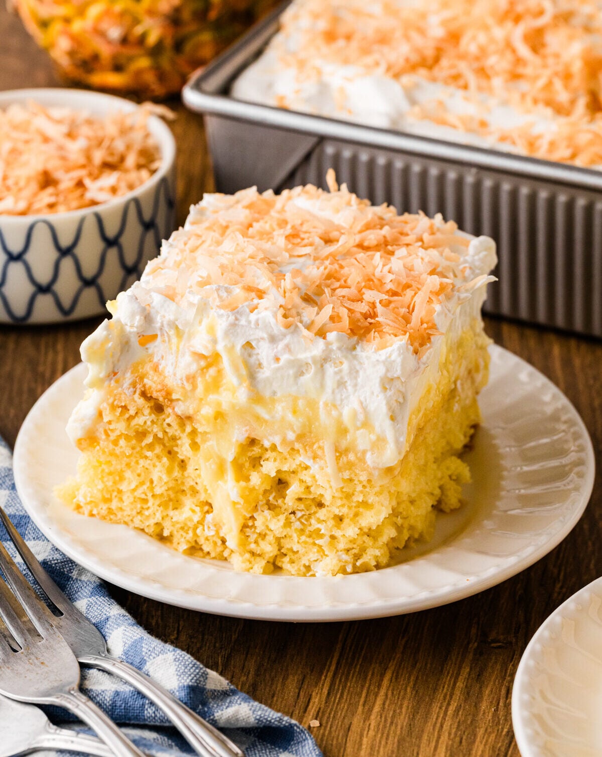 Pineapple poke cake slice topped with whipped cream and toasted coconut flakes on a white plate with a fork