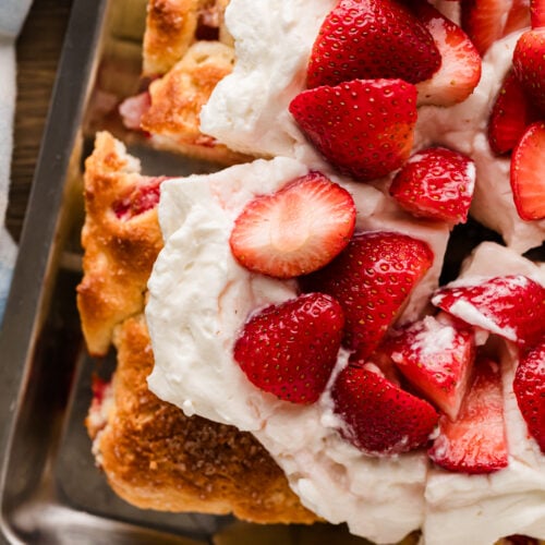 Strawberry Shortcake Sheet Cake in a metal baking pan topped with whipped cream and fresh sliced strawberries, sliced and ready to serve