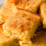Golden-brown sour cream cornbread squares with crispy edges stacked on a white plate, showing tender crumb texture