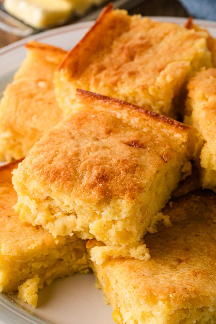 Squares of sour cream cornbread stacked on a white plate with golden edges and a soft, buttery crumb.