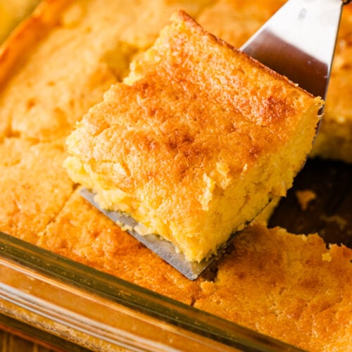 Golden sour cream cornbread in glass baking dish with one square piece lifted on spatula, showing moist crumb texture