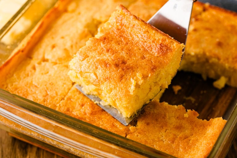Golden sour cream cornbread in glass baking dish with one square piece lifted on spatula, showing moist crumb texture