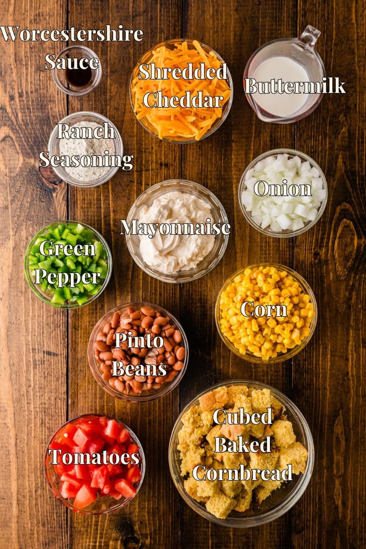 Overhead flat-lay of labeled ingredients for Southern Cornbread Salad in glass bowls — cubed baked cornbread, diced tomatoes, pinto beans, sweet corn, diced green bell pepper, chopped onion, mayonnaise, ranch seasoning, shredded cheddar cheese, buttermilk and Worcestershire sauce arranged on a dark wood surface